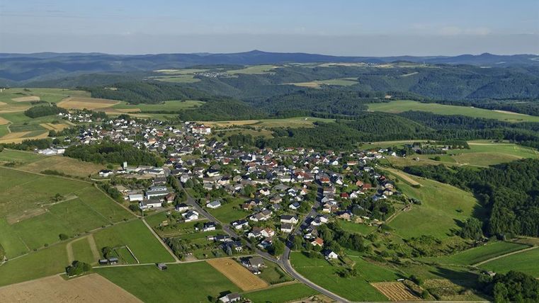 Eine malerische Landschaft mit einem kleinen Dorf, umgeben von sanften Hügeln und Feldern. Der klare Himmel ergänzt das idyllische Panorama.